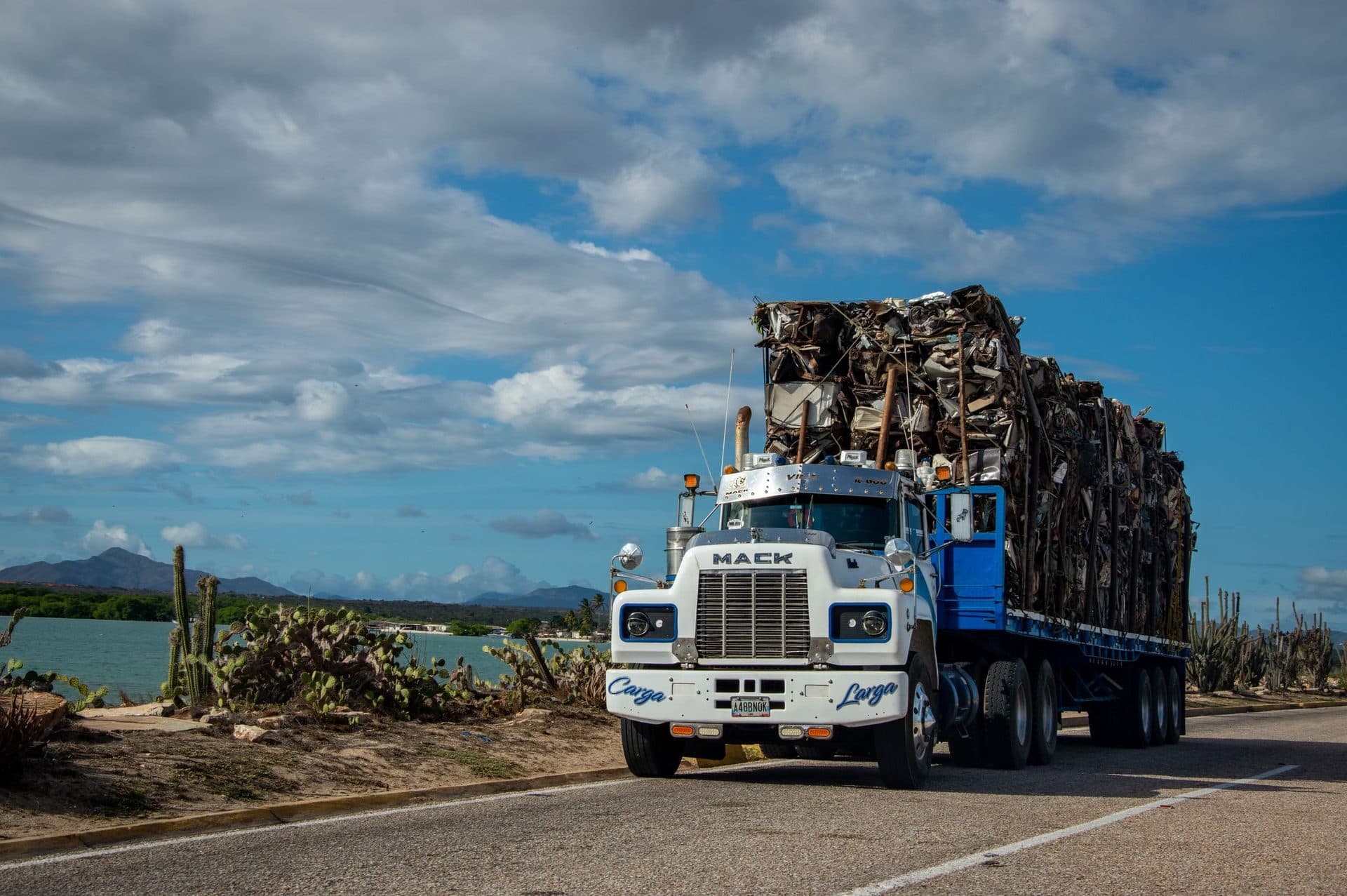 White Mack tractor hauling a 53-foot flatbed loaded with baled scrap metal on a coastal road