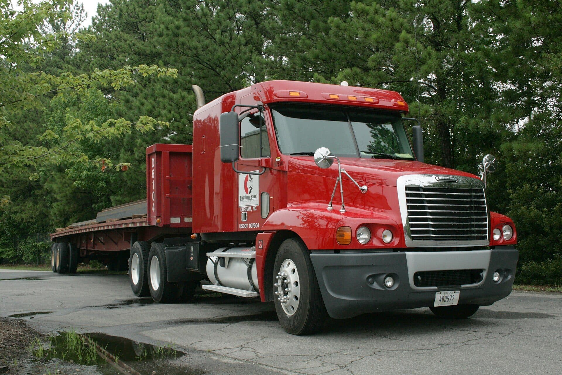 Red Freightliner tractor with an empty 53-foot flatbed parked by trees