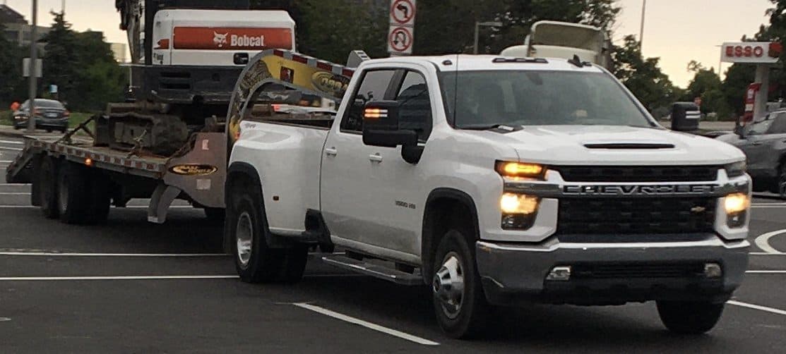 White heavy-duty pickup pulling a gooseneck flatbed carrying equipment at an intersection