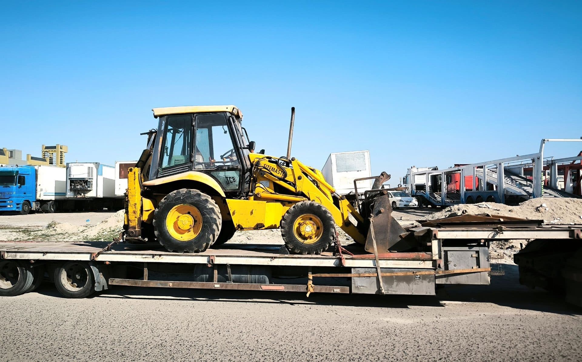Yellow backhoe loader secured on a flatbed trailer in an industrial yard